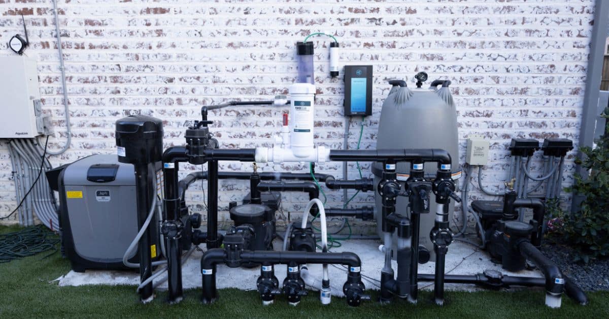A backyard pool equipment pad with a typical pool pump and filter next to a breaker panel in the background, photographed in natural daylight