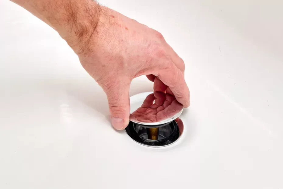A bathroom photo showing a hand turning a chrome toe-touch bathtub stopper counterclockwise while a towel protects the tub