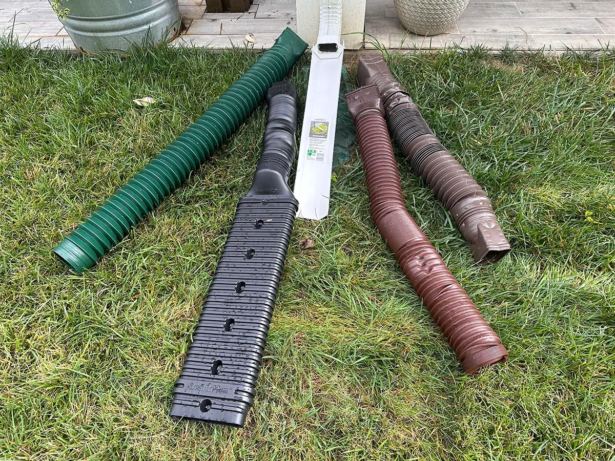 A black flexible downspout extension connected to a gutter downspout and discharging water several feet away from a house foundation, real photo