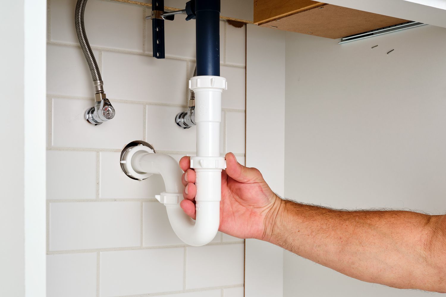 A bucket positioned under a kitchen sink P-trap while a homeowner loosens plastic slip nuts by hand, real photograph