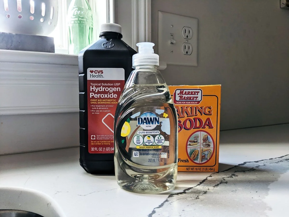 A bucket with an open bottle of 3% hydrogen peroxide, a box of baking soda, and a small bottle of dish soap next to nitrile gloves
