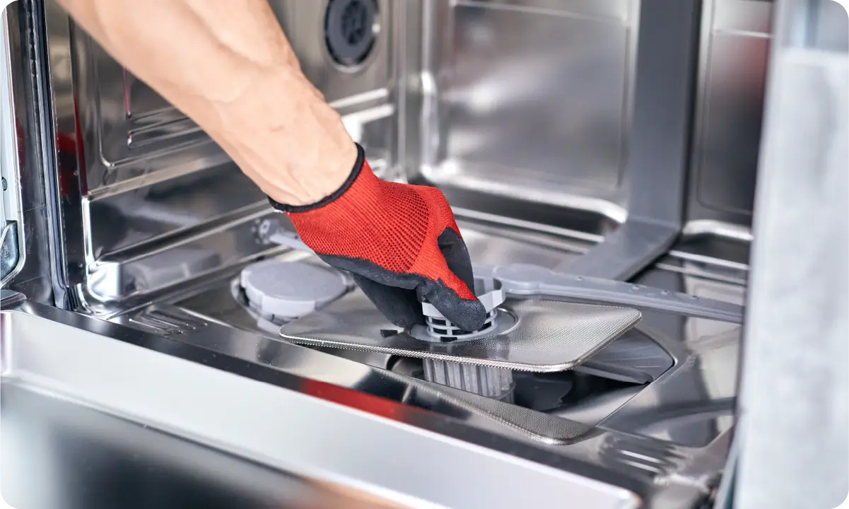 A built-in stainless steel dishwasher in a home kitchen with the door cracked open and a homeowner kneeling nearby holding a flashlight, realistic indoor photo