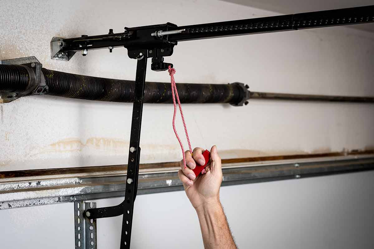 A ceiling-mounted garage door opener with a red emergency release handle hanging from the trolley on the opener rail
