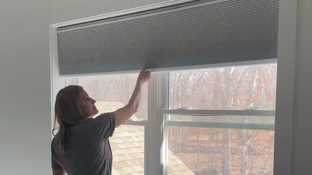 A cellular shade headrail being lifted into mounting brackets inside a window frame, showing the headrail engaging the bracket lip