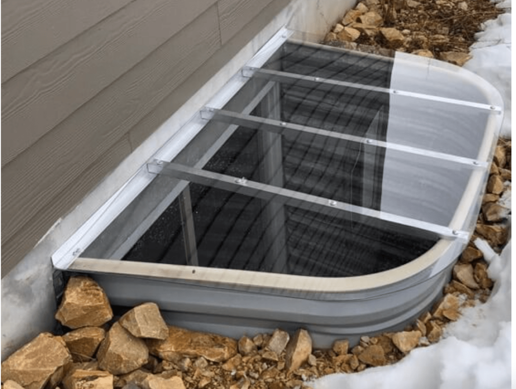 A clear bubble-style polycarbonate window well cover secured over a galvanized metal window well beside a house foundation, real photo