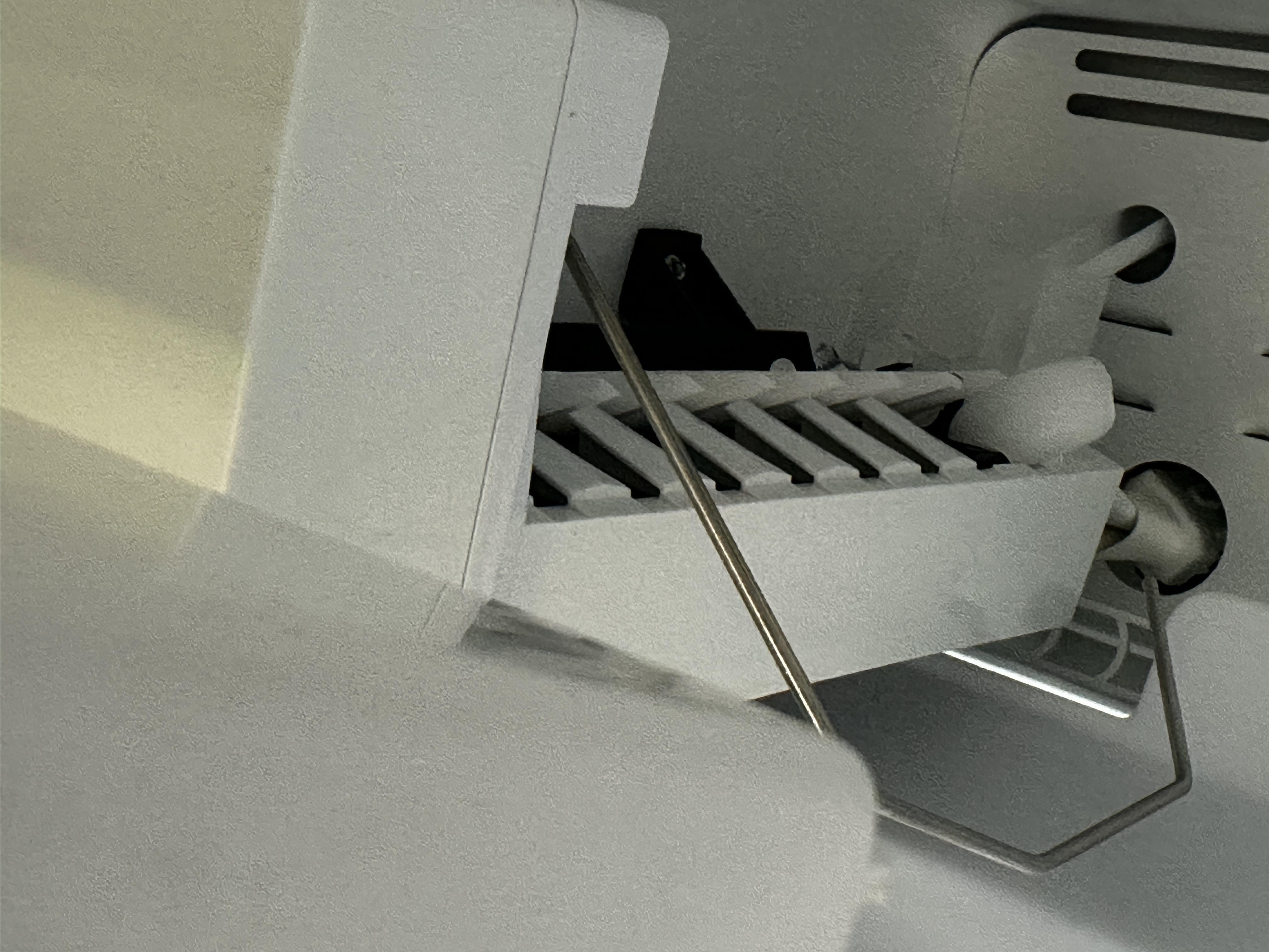 A close-up inside a freezer showing an ice maker ejector area with a cube wedged near the rotating ejector fingers