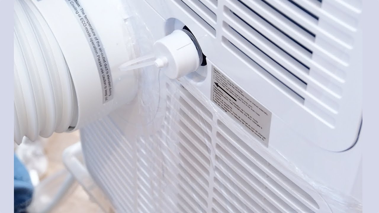 A close-up of a portable air conditioner lower drain port with the plug removed and water draining into a shallow tray