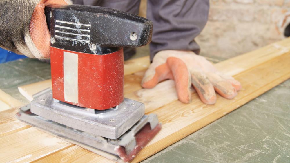 A close-up of a sheet sander with a rectangular pad being used on a flat wooden panel