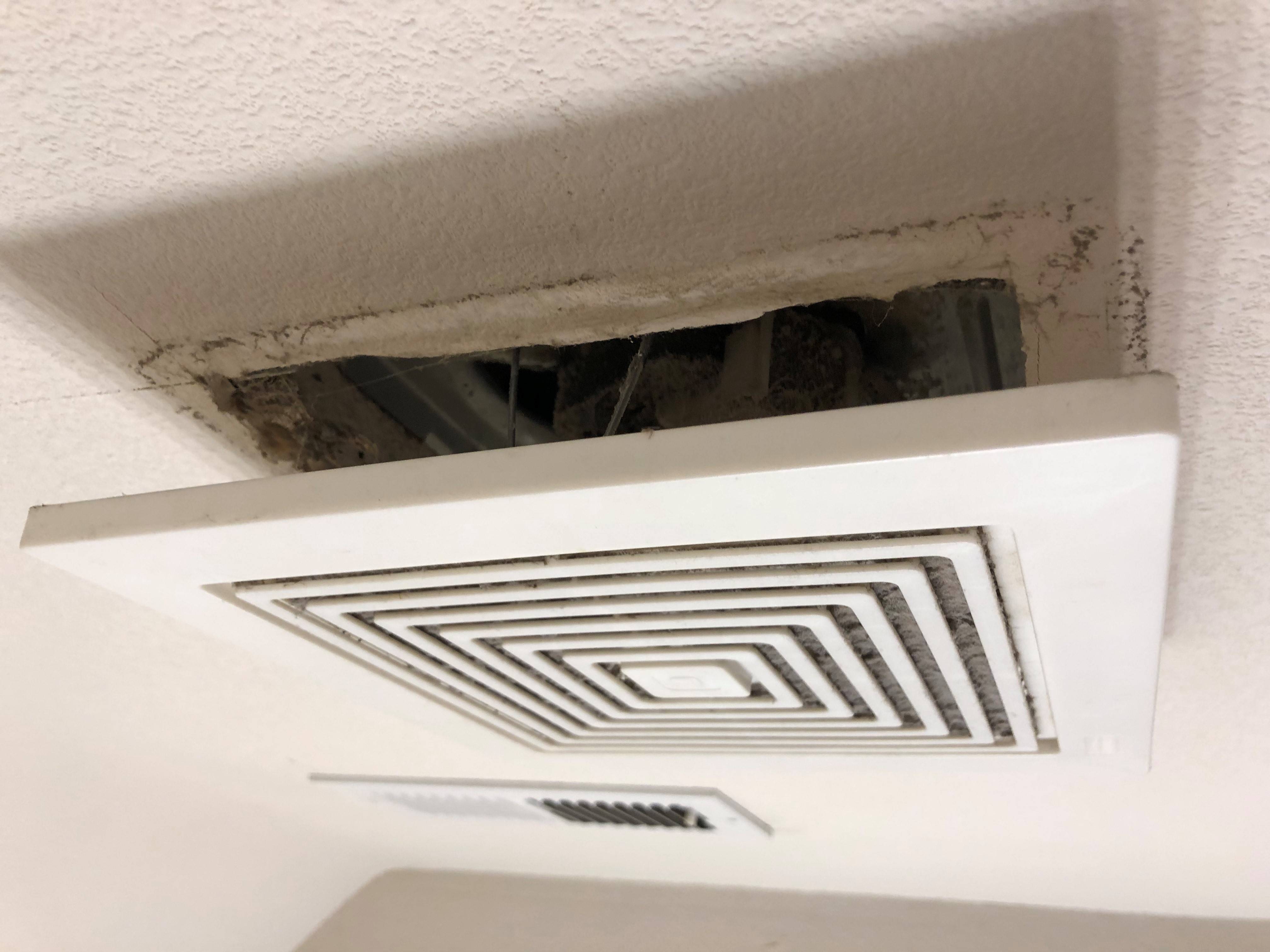 A close-up photo of a bathroom exhaust fan in the ceiling with the plastic grille pulled down, showing the spring clips and dusty fan housing edge, realistic indoor lighting