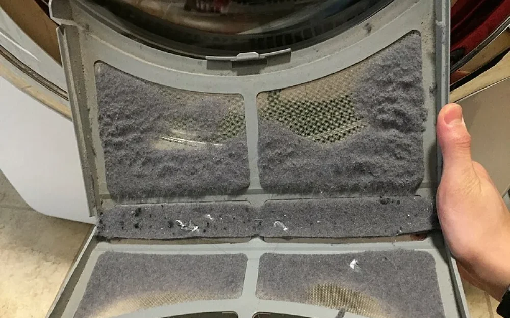 A close-up photo of a dryer door latch area with visible lint buildup being removed using a vacuum crevice tool