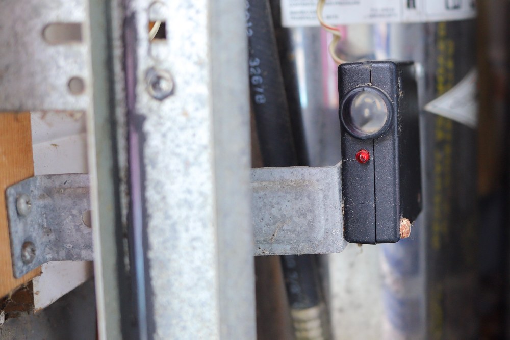 A close-up photo of a garage door safety sensor mounted near the floor on a metal track, with the small lens facing across the doorway toward the matching sensor
