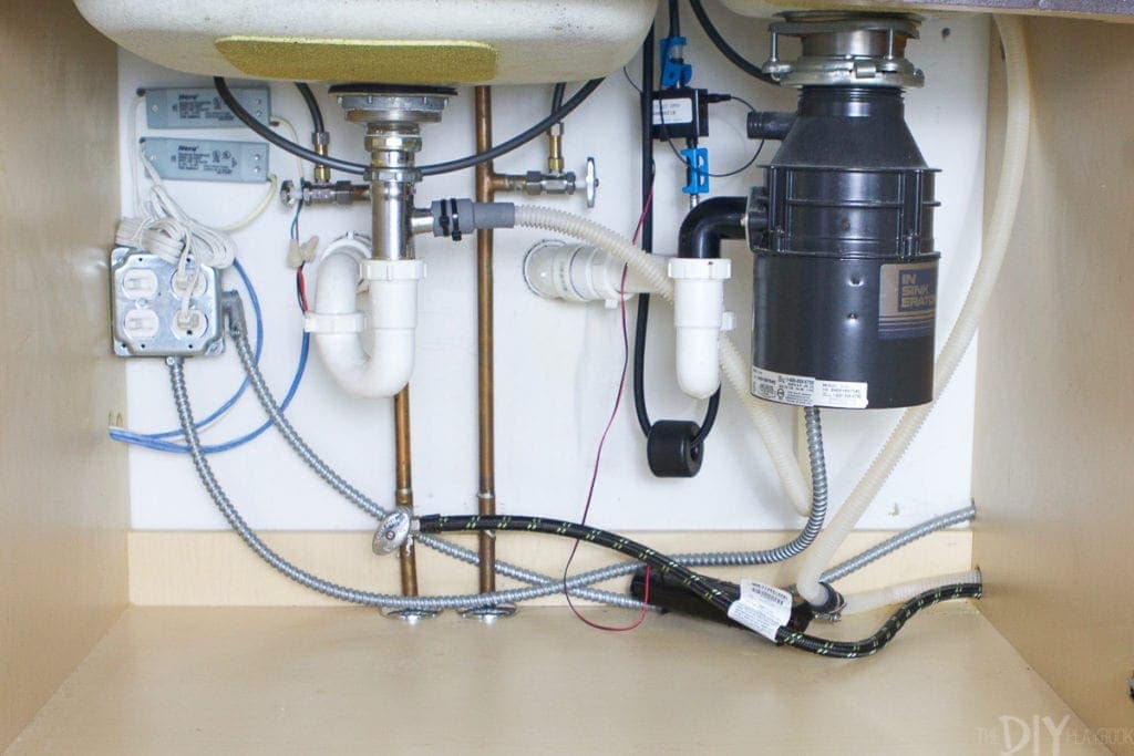 A close-up photo of a garbage disposal power cord unplugged from an under-sink outlet, with a bucket and towels nearby