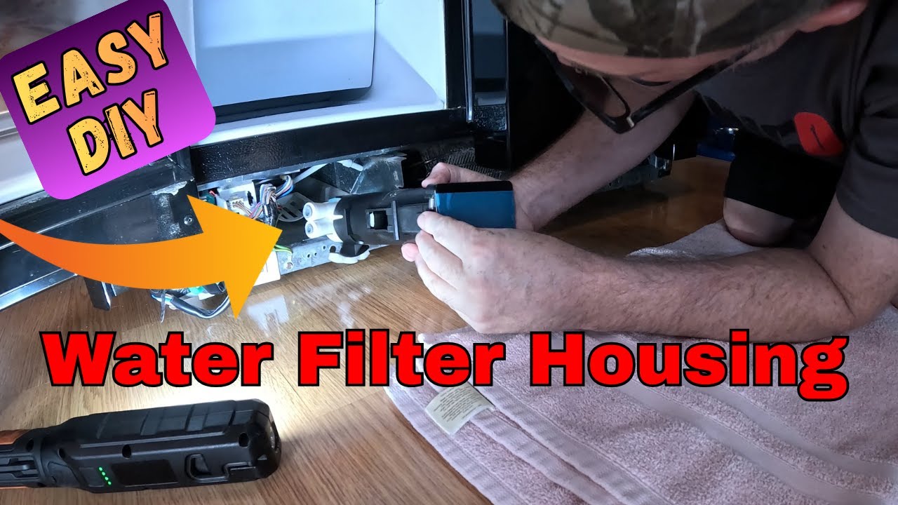 A close-up photo of a hand holding a folded paper towel under a refrigerator water filter housing while inspecting for moisture