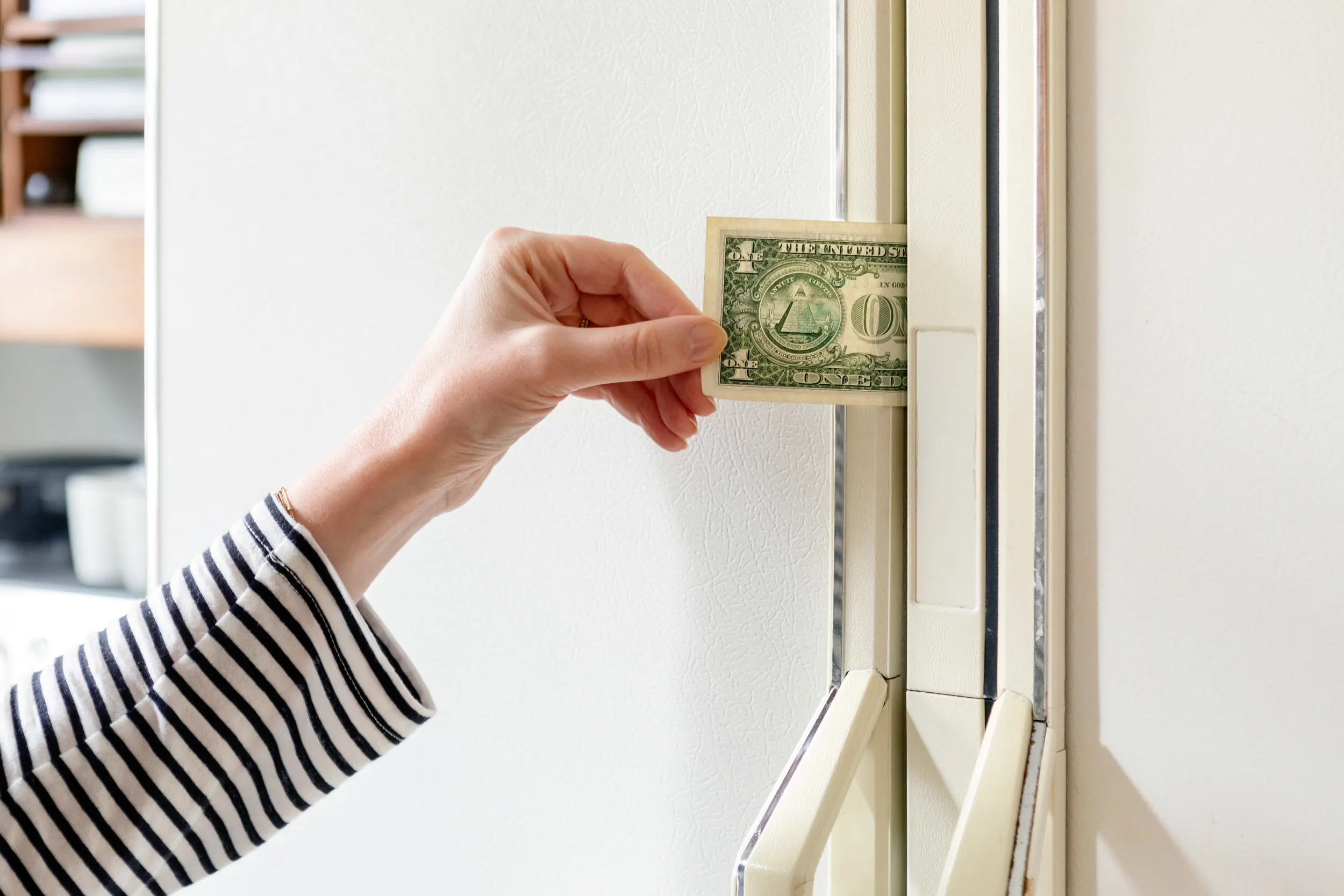 A close-up photo of a hand pulling a dollar bill from a refrigerator door seal while the door is mostly closed, showing a common gasket leak test