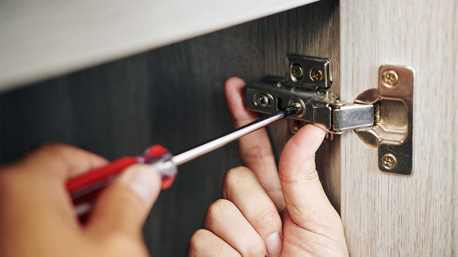 A close-up photo of a kitchen cabinet door hinge being adjusted with a screwdriver inside the cabinet, natural indoor lighting