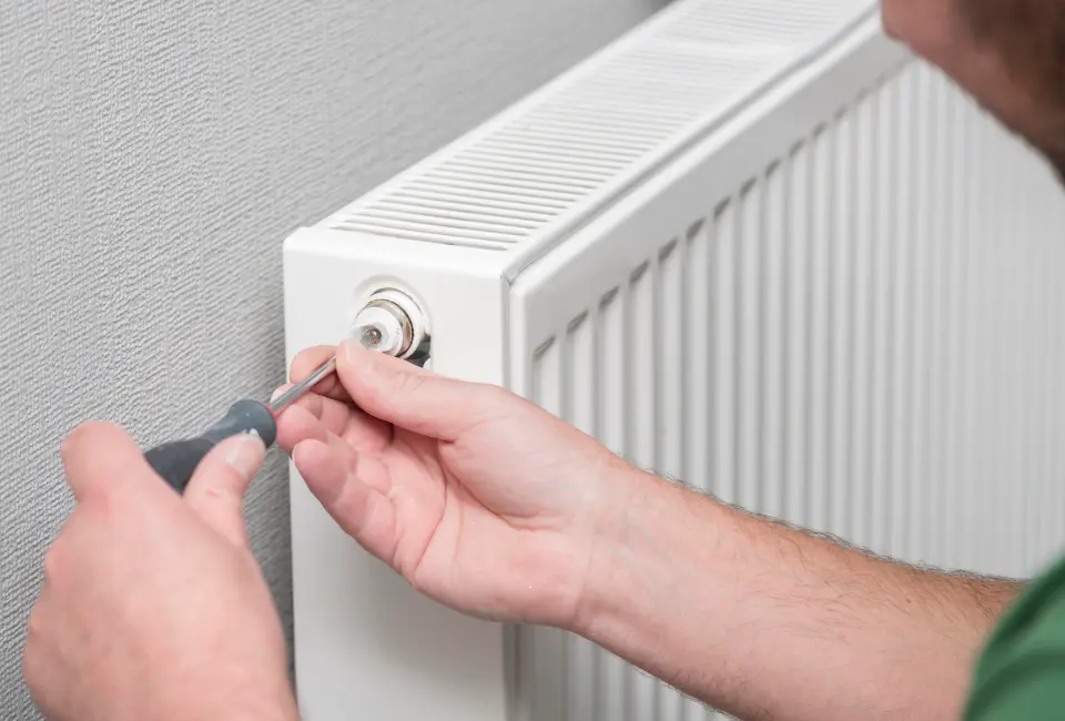 A close-up photo of a person using a radiator key to bleed a white panel radiator while holding a small cup under the bleed valve