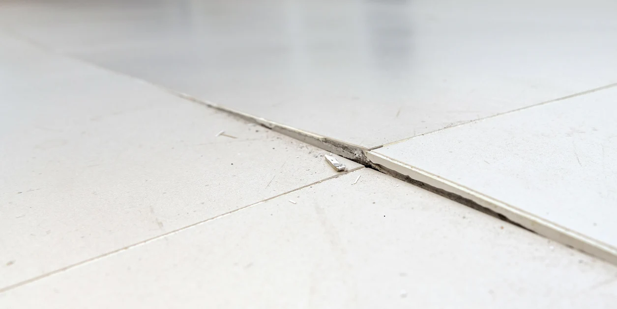 A close-up photo of a single cracked ceramic floor tile in a kitchen floor, with surrounding intact tiles and visible grout lines, natural window light, realistic home renovation photography
