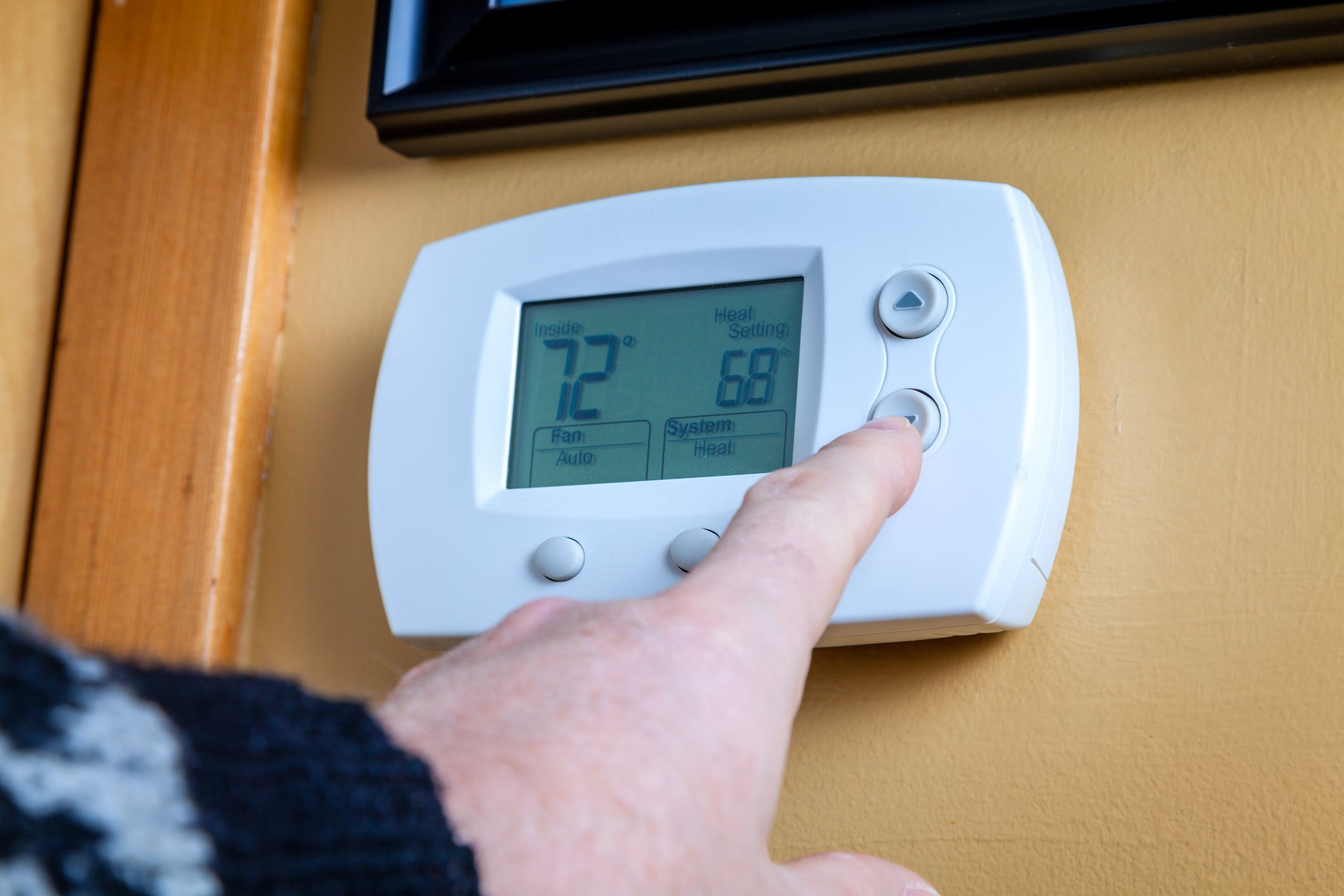A close-up photo of a standard digital thermostat mounted on a light-colored interior wall in a lived-in home hallway, with a homeowner's hand reaching toward the buttons, natural window light