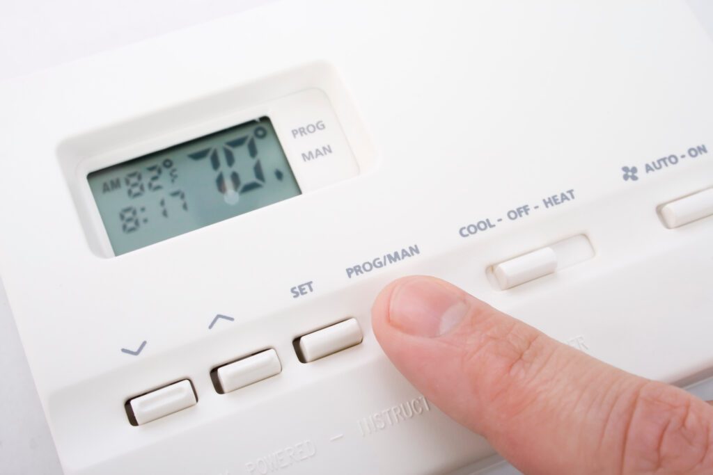 A close-up photo of a wall thermostat displaying AUX heat while a hand hovers near the mode button in a lived-in hallway