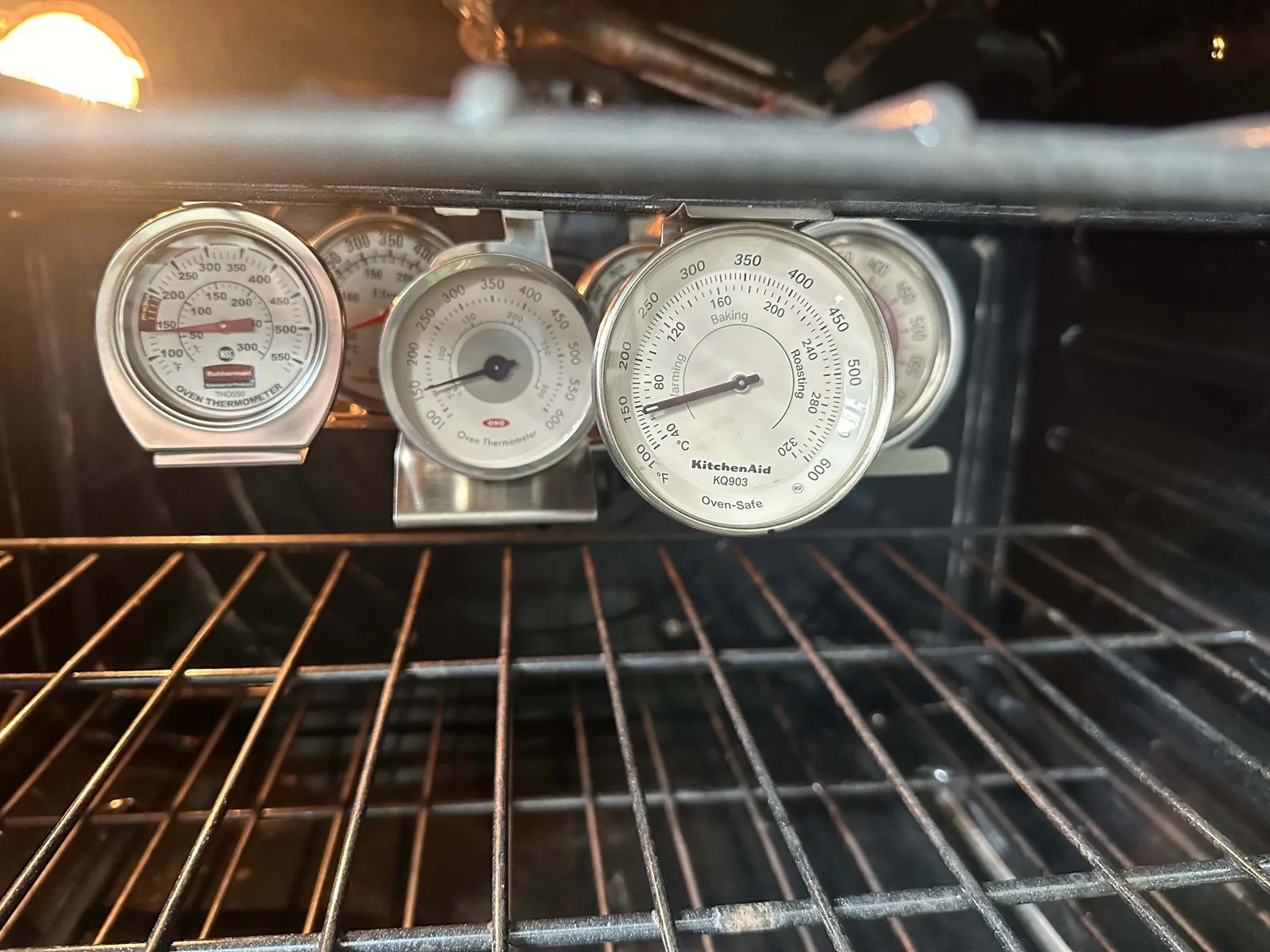 A close-up photo of an analog oven thermometer standing on the middle rack inside a lit oven cavity during preheating