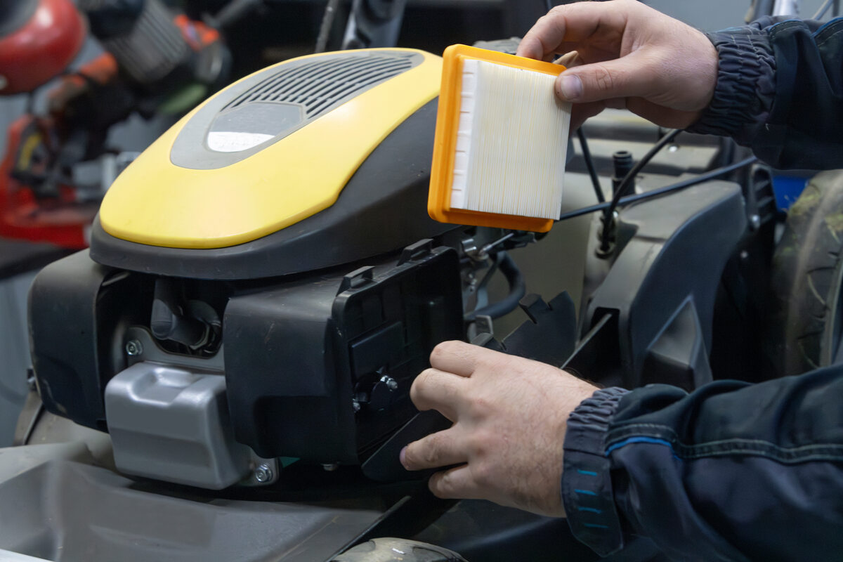 A close-up photo of hands opening a lawn mower air filter cover to remove a dusty foam air filter