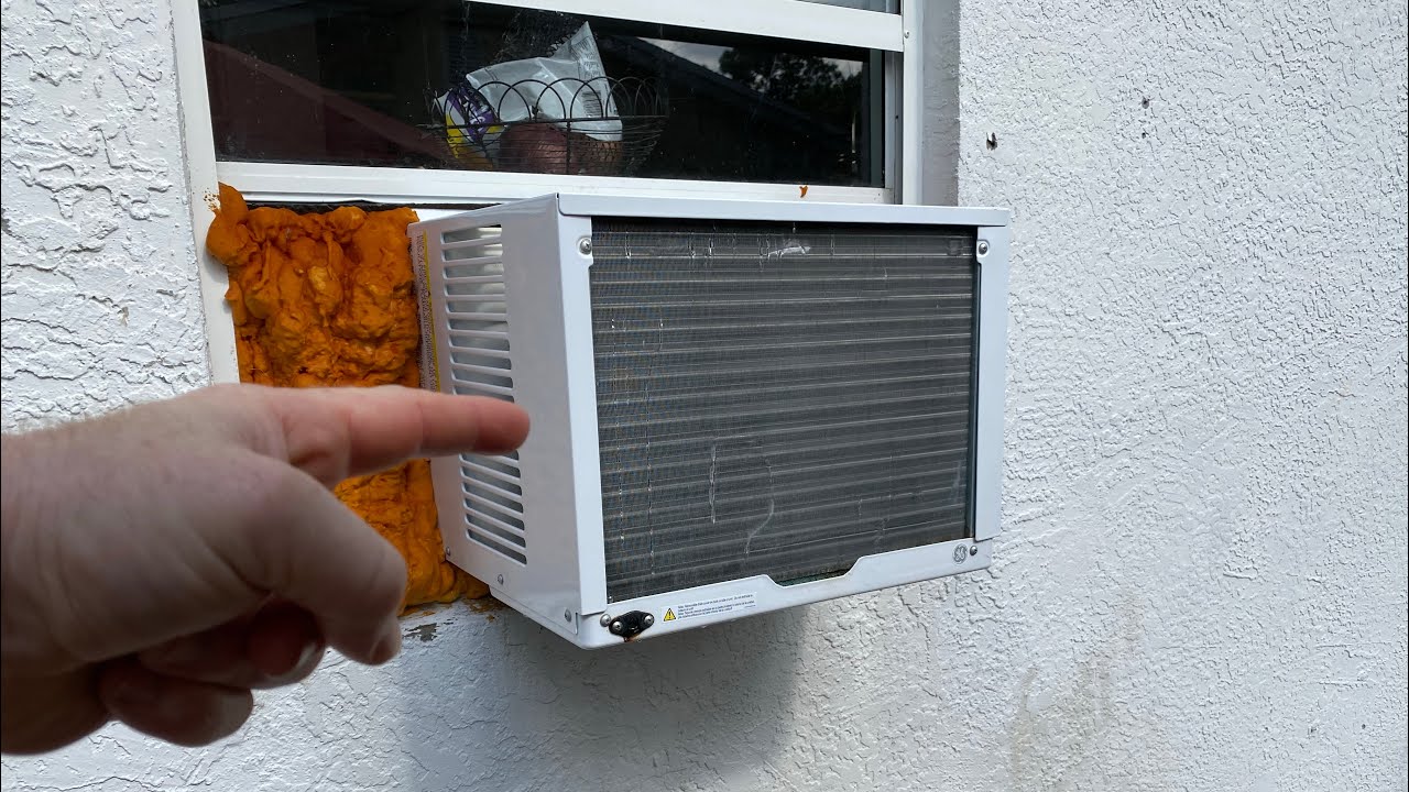 A close-up photo of the outdoor side of a window air conditioner with the cover removed, showing a small leaf lodged near the fan blade and a gloved hand reaching in to remove it