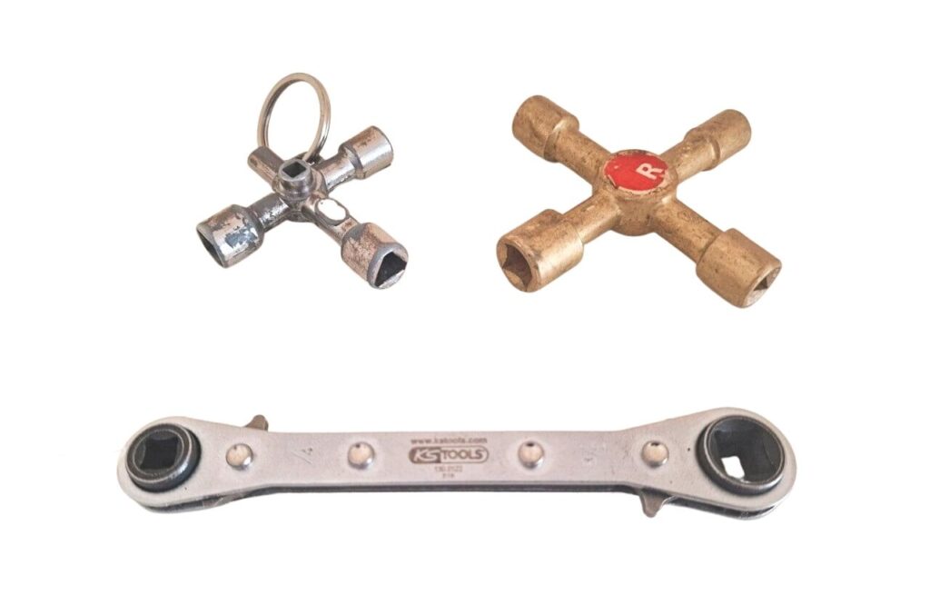 A close-up photograph of a person holding a small radiator bleed key against a brass bleeder valve on the side of a painted cast-iron radiator, with a towel and small cup positioned underneath