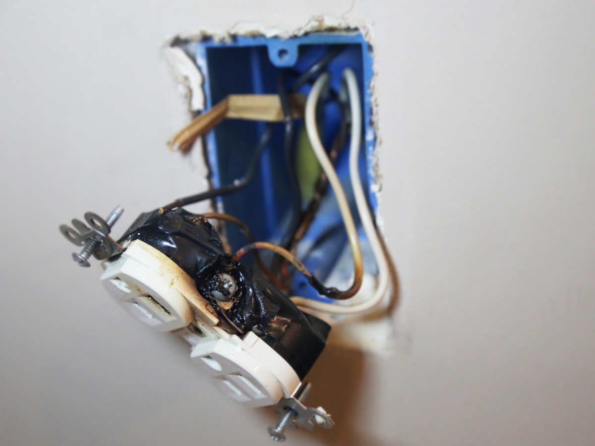 A close-up photograph of an electrical outlet pulled from a wall box showing wires inserted into the push-in backstab holes