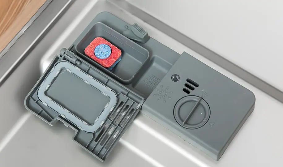 A close-up real photo of a dishwasher door interior with the detergent dispenser door open, showing the hinge and latch area