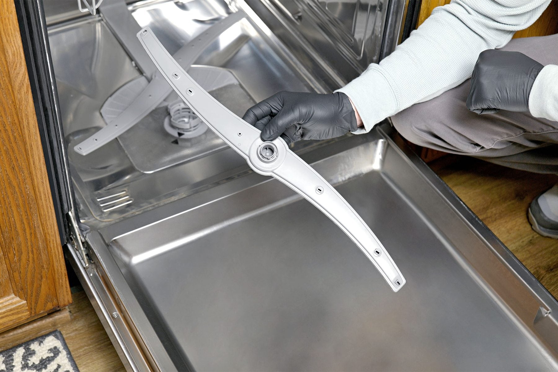 A close-up real photo of a dishwasher spray arm being rinsed in a sink while a hand clears clogged jet holes with a toothpick