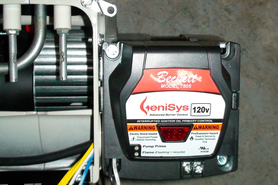 A close-up real photo of an oil burner primary control box mounted on the burner, with a visible reset button and indicator light