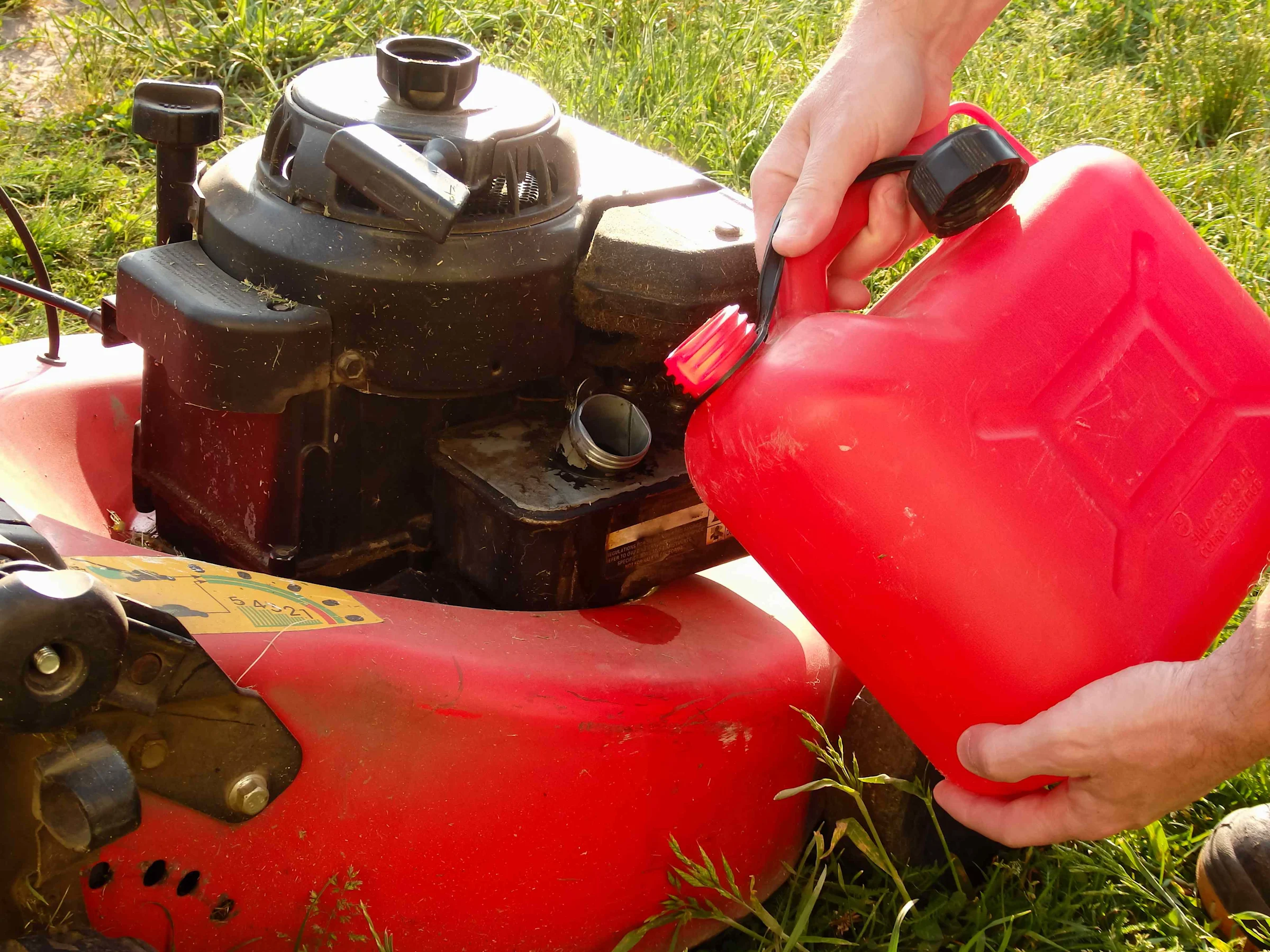 A close-up real photo of fresh gasoline being poured from a red gas can into a small engine fuel tank outdoors