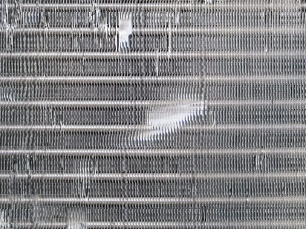 A close-up view through a portable air conditioner intake showing dusty metal coil fins and the edge of a removable filter frame