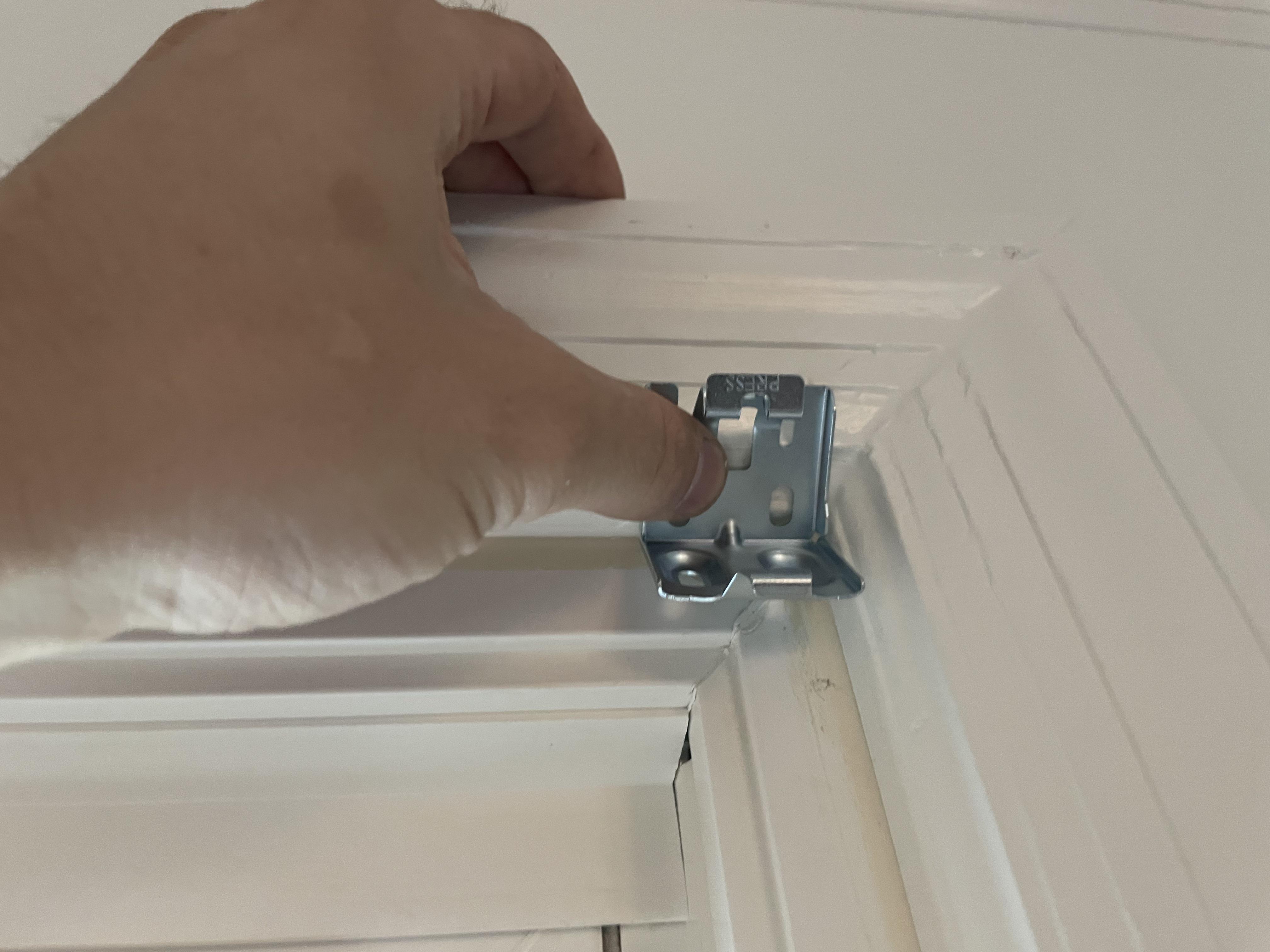 A close view of a drill driving a screw through a window blind mounting bracket into wooden trim