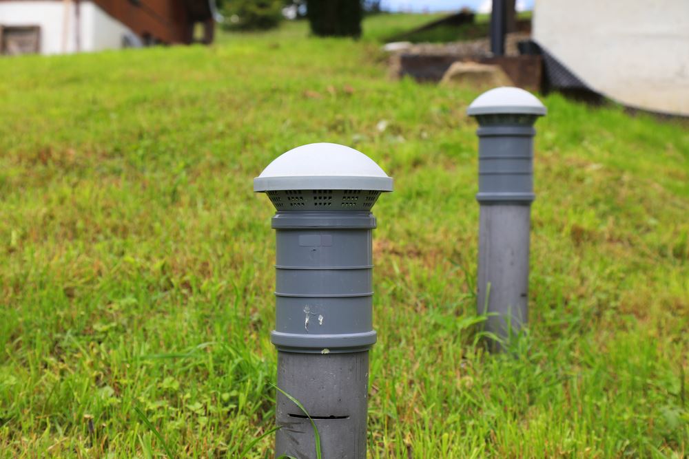 A close view of a small PVC vent pipe near a house foundation in a residential yard, with a homeowner standing back a few feet, realistic photo