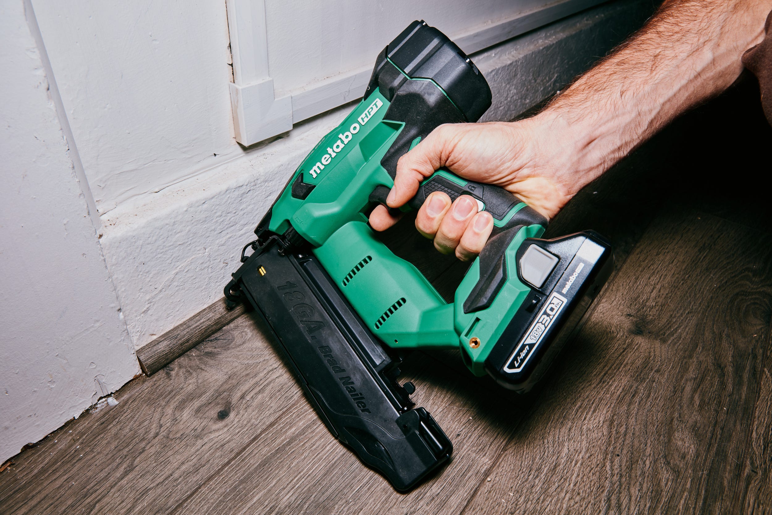 A cordless brad nailer being used to fasten white baseboard trim to a painted wall in a home renovation setting, natural light, realistic photography