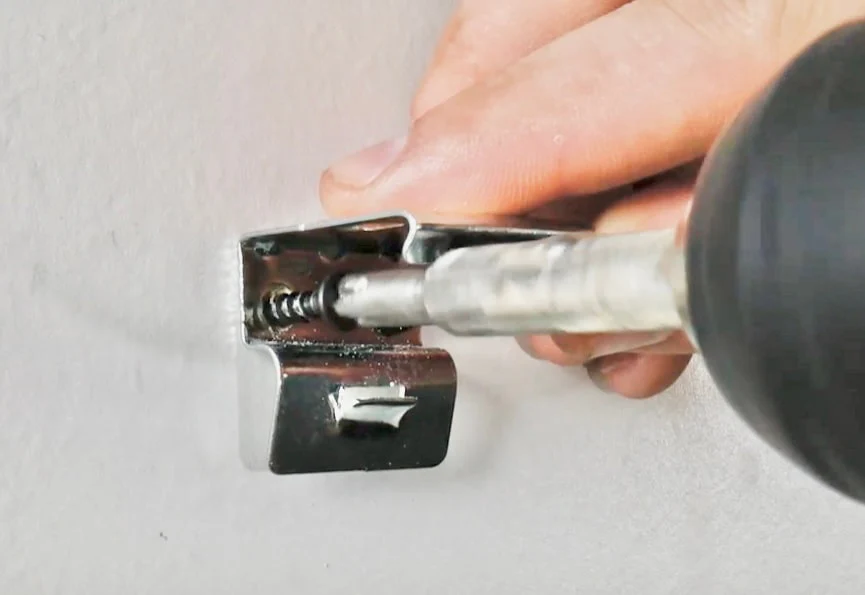 A drill driving a screw through a shade bracket into a wooden window frame