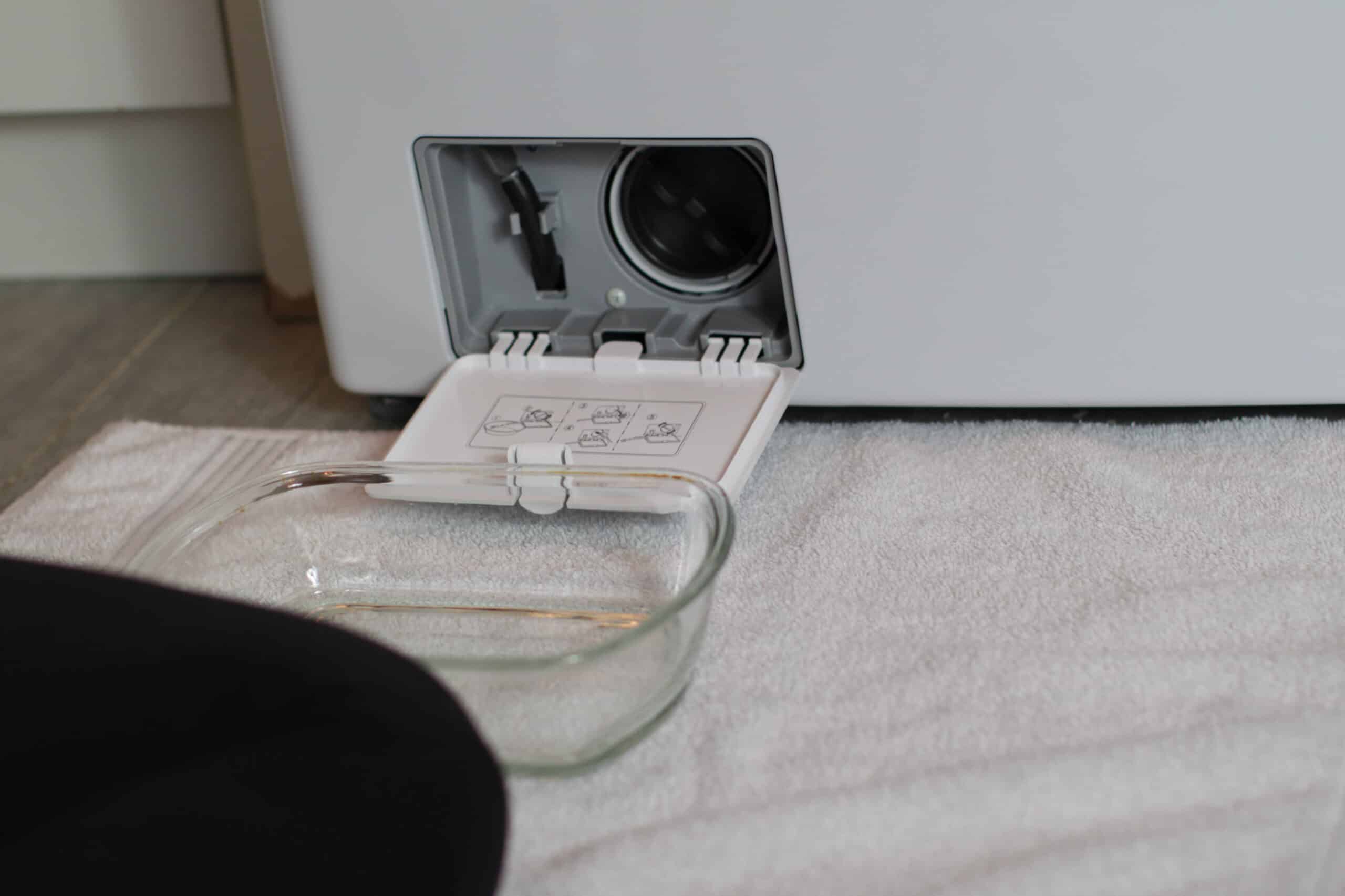 A front-loading washing machine with the lower access panel open, showing a hand loosening the round drain pump filter cap