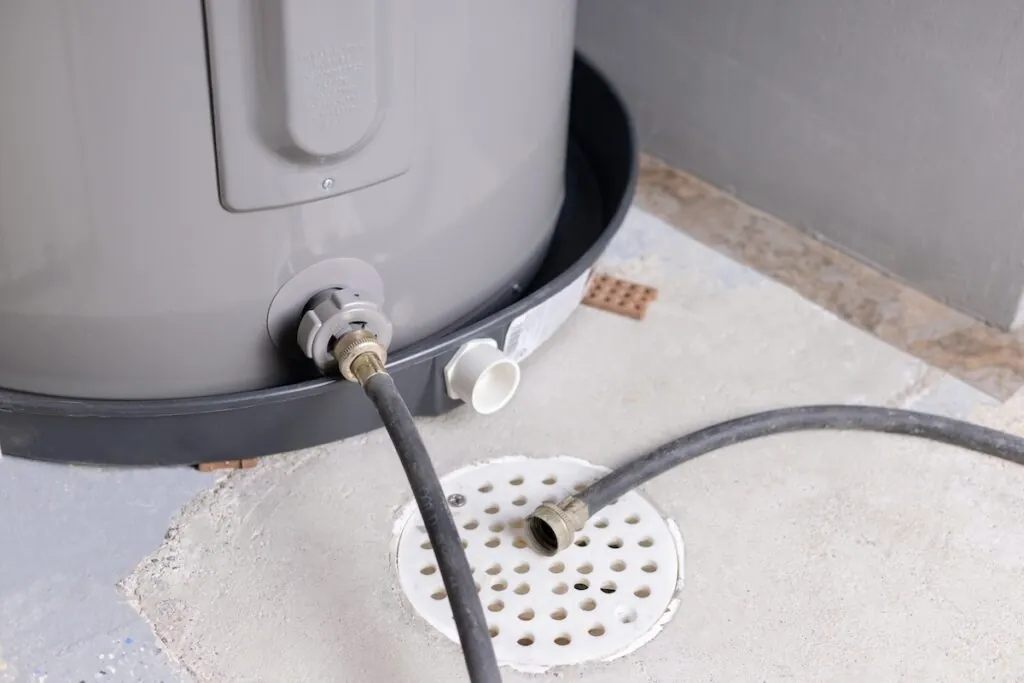 A garden hose connected to an electric water heater drain valve with water flowing into a floor drain in a utility room, realistic photo