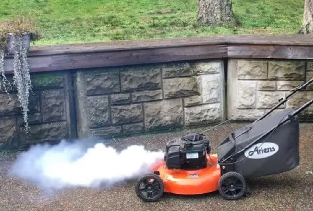 A gas-powered push lawn mower on a driveway with a small plume of white smoke rising from the engine area in natural daylight