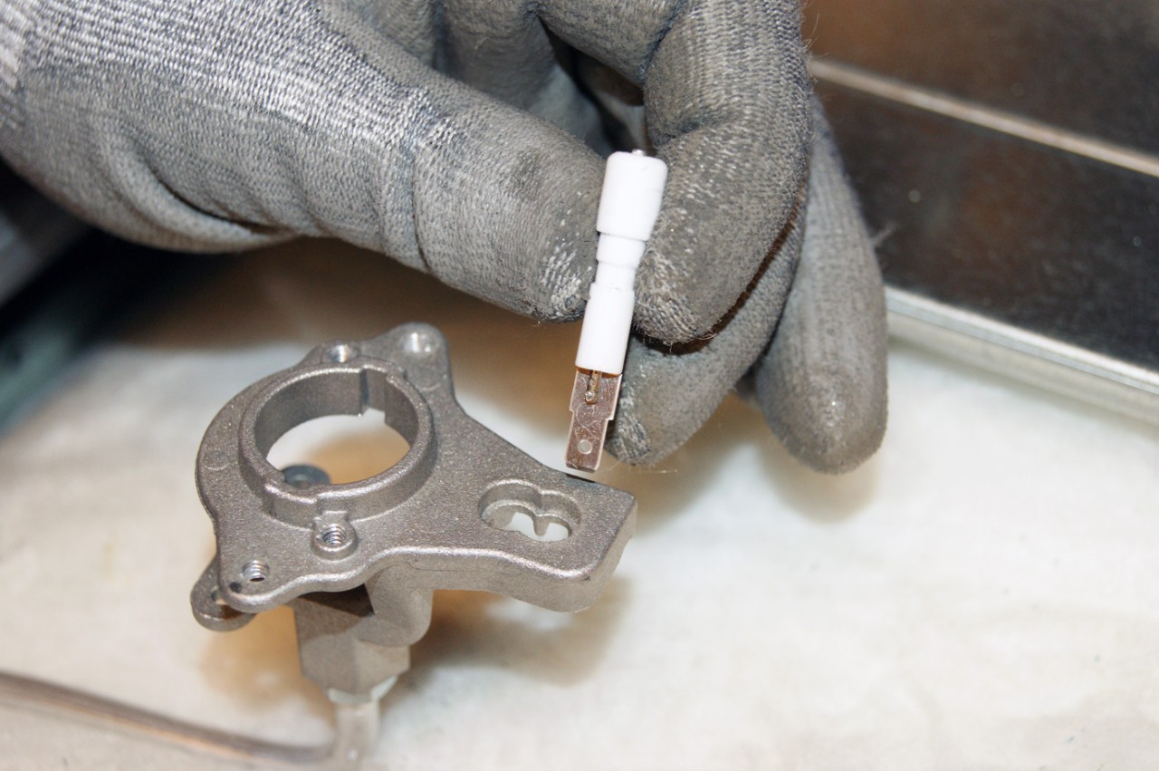 A gas stove spark igniter electrode next to a burner head, showing the small ceramic insulator and metal tip area that can stay damp after cleaning