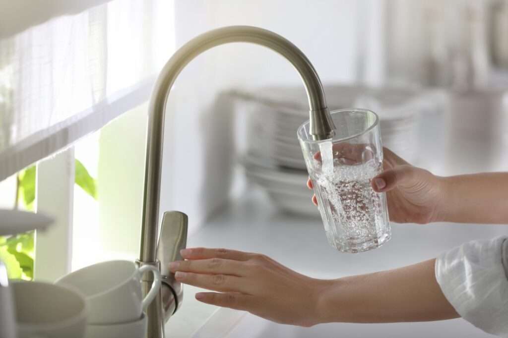 A hand holding a clear drinking glass filled with tap water near a kitchen sink, realistic indoor household photo