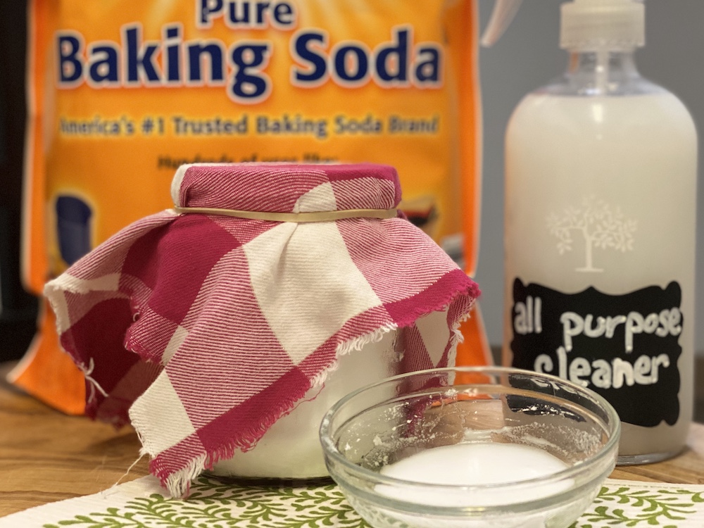 A hand wiping the inside wall of a refrigerator using a microfiber cloth and a bowl of baking soda cleaning solution on the nearby counter