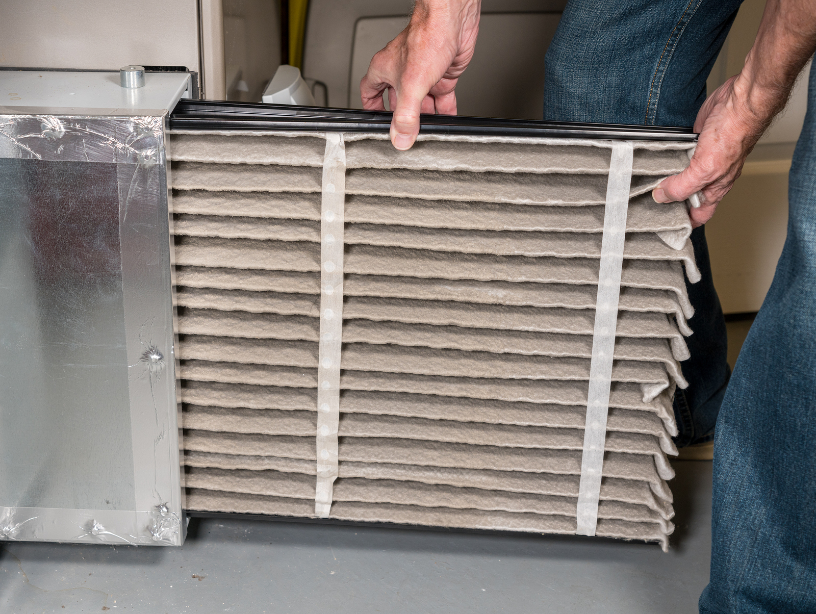 A homeowner holding a pleated furnace air filter in front of a return air filter slot on a furnace, hands visible, indoor basement setting, photorealistic
