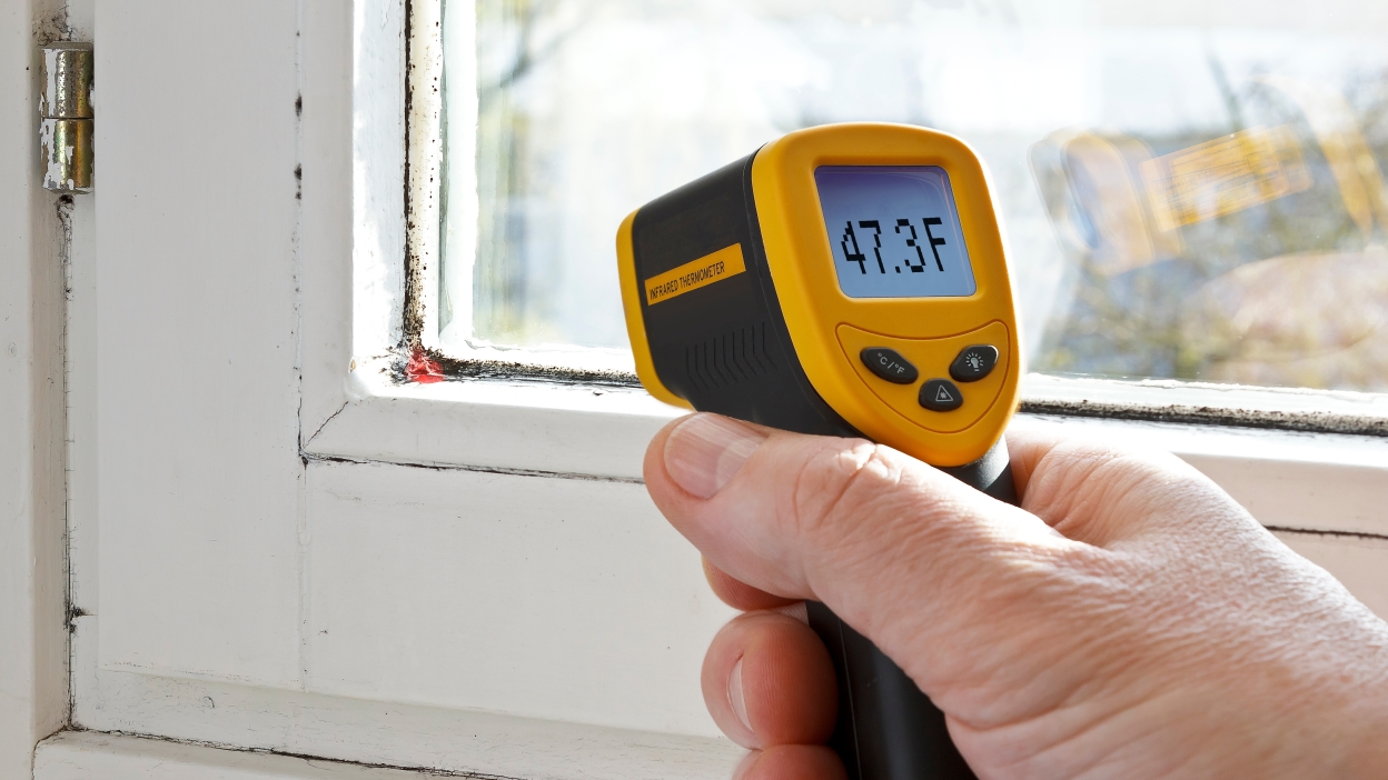 A homeowner holding an infrared thermometer aimed at a wall outlet cover plate in a well-lit room, realistic home photo