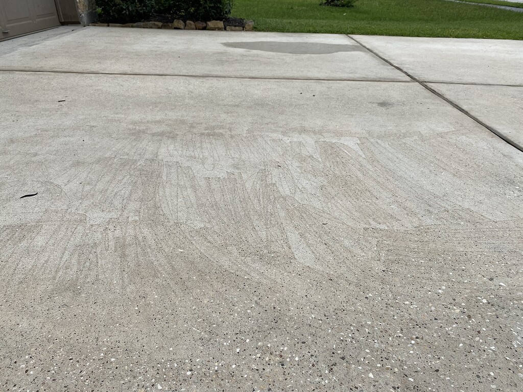 A homeowner kneeling beside a gas pressure washer on a driveway with the garden hose connected and the high-pressure hose laid out, checking fittings and the pump area in natural daylight