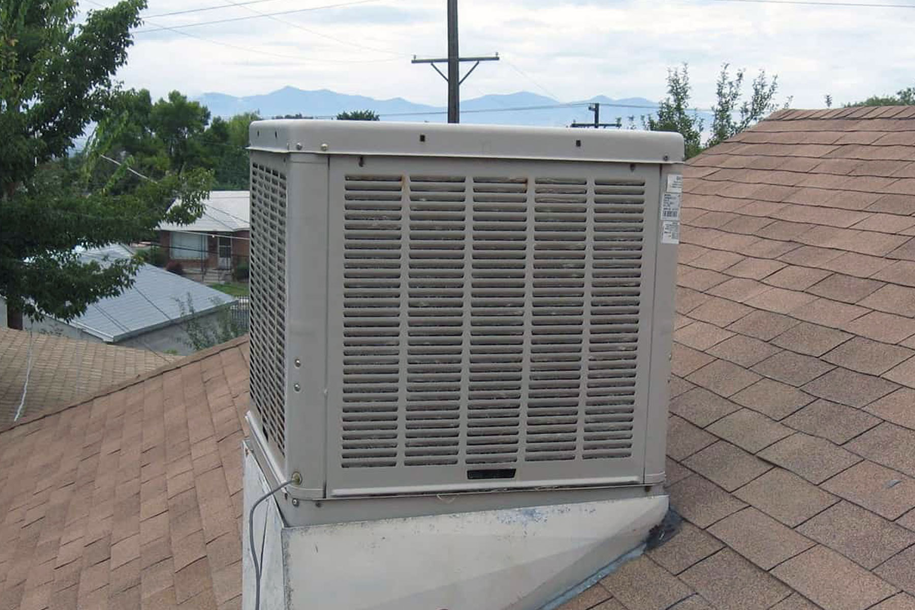 A homeowner kneeling beside a roof-mounted evaporative cooler with the access panel open, checking wet pads and water lines in bright daylight