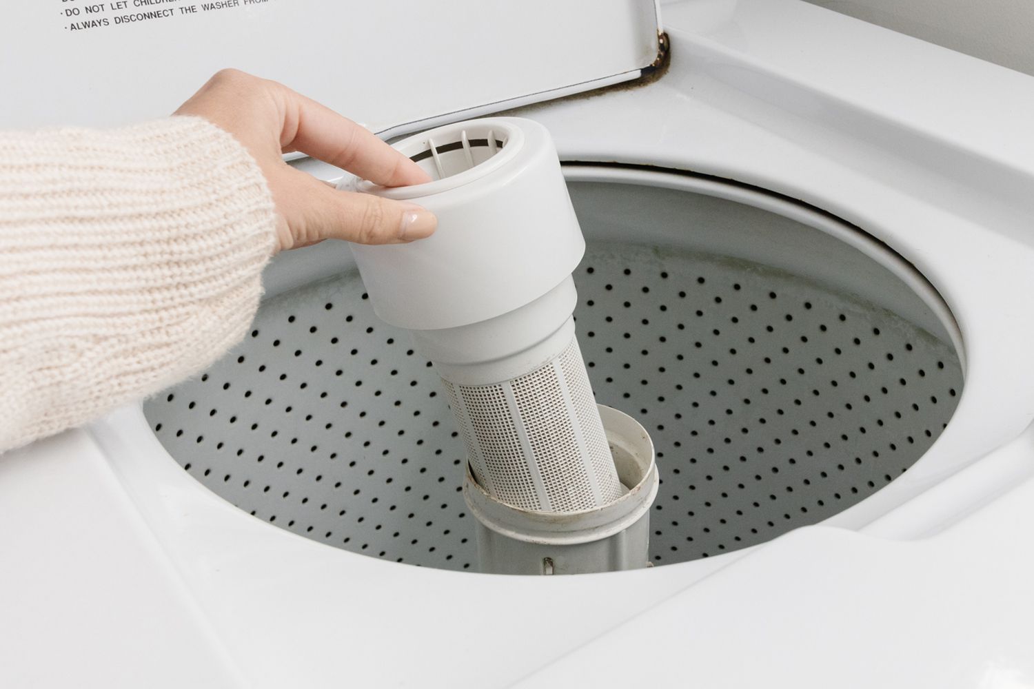 A homeowner kneeling beside a top-load washing machine with a shallow pan and towels on the floor, preparing to open a small access panel near the bottom of the washer