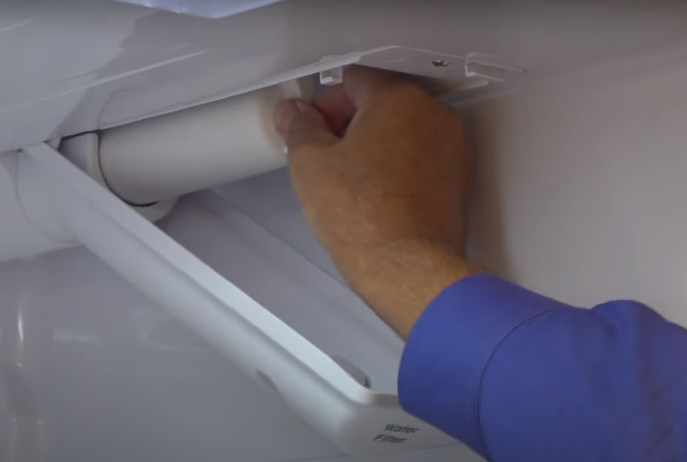 A homeowner kneeling in front of a stainless steel refrigerator with the lower grille open, hands positioned on a water filter housing, indoor kitchen photo