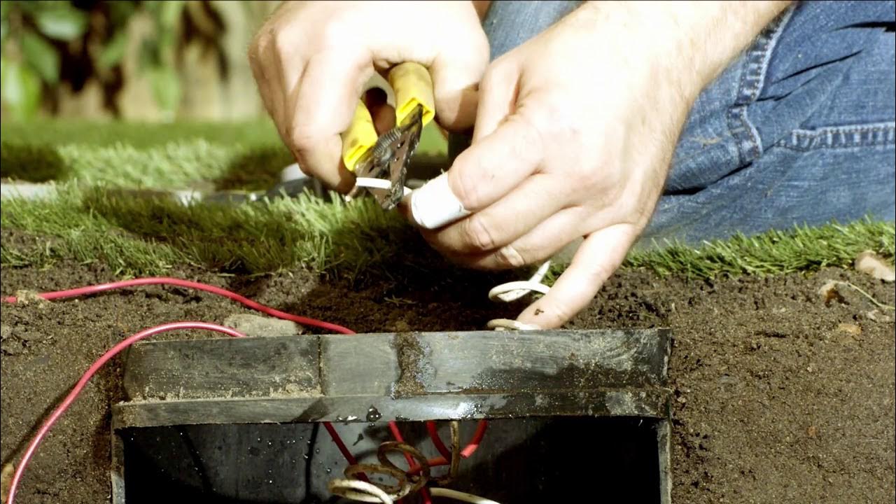 A homeowner kneeling next to an open sprinkler valve box, hand touching a solenoid on a plastic irrigation valve, daylight photo